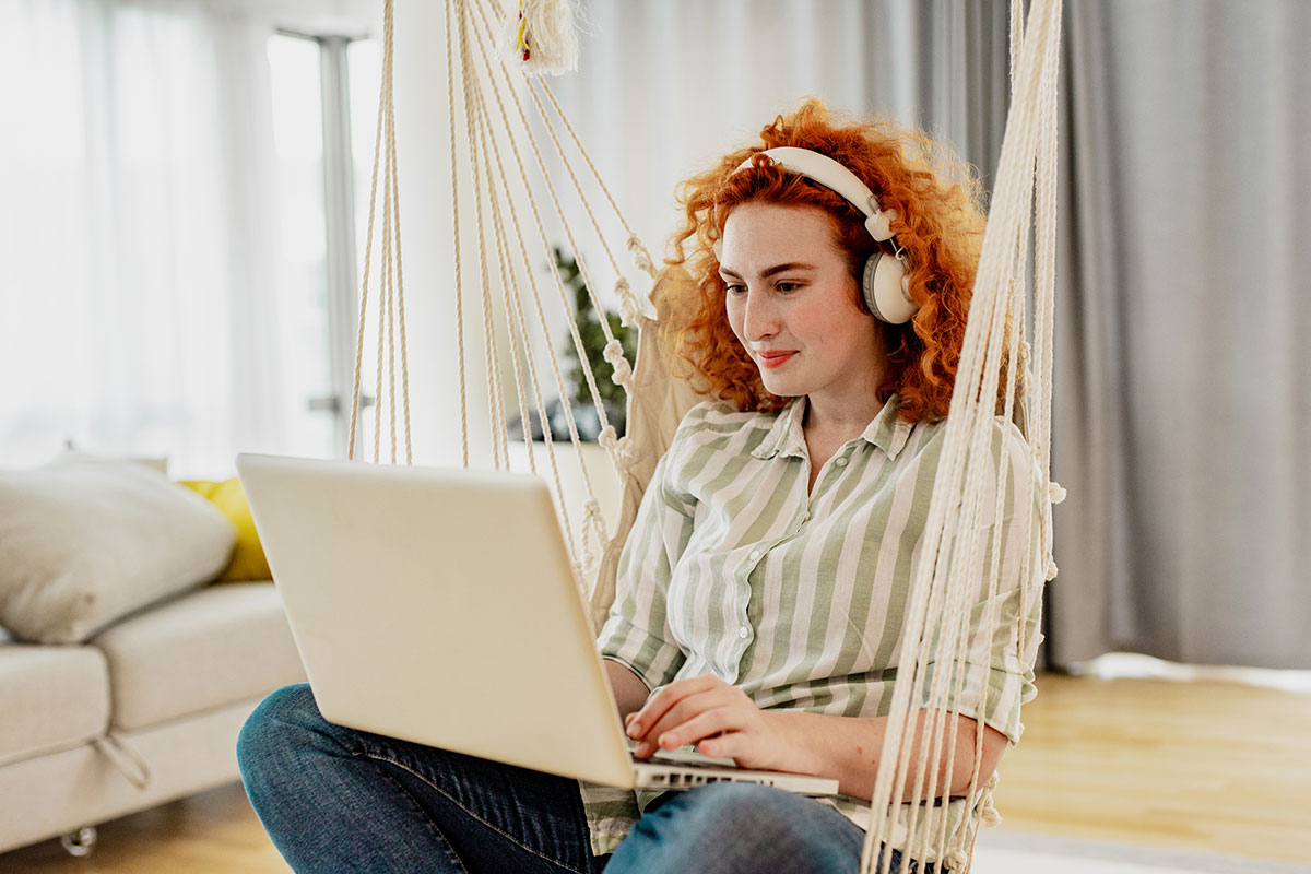 Frau mit Kopfhörern sitzt in einer Hängematte und benutzt einen Laptop in einem hellen Raum.